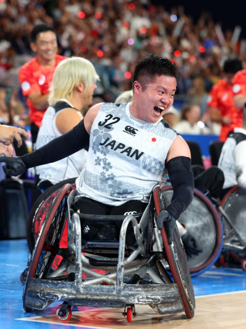 A man in a wheelchair yells happily. He is on a wheelchair rugby court. More people in wheelchairs behind him smile, the crowd in the stands cheers.