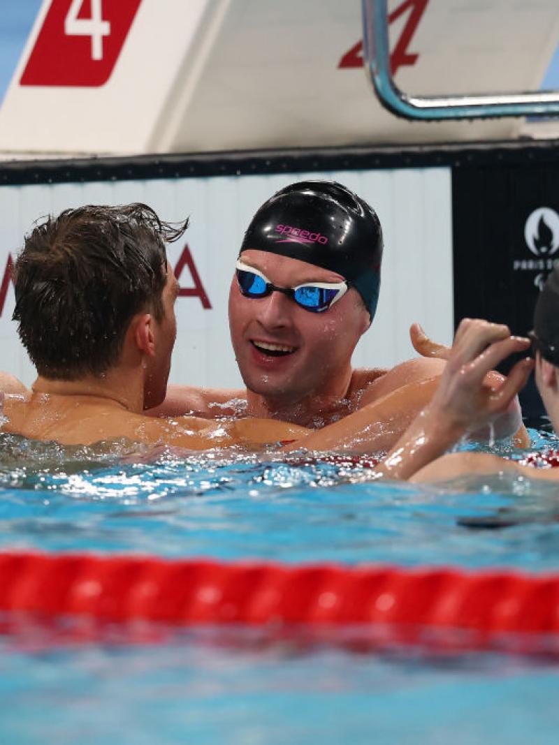 Three men are in a swimming pool, two are congratulating a third, who is wearing a swim cap and goggles.