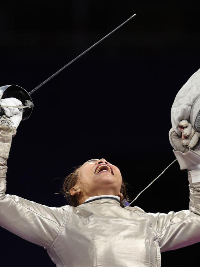 A woman competing in wheelchair fencing holds up her sabre in one hand and her face shield in the other and yells passionately