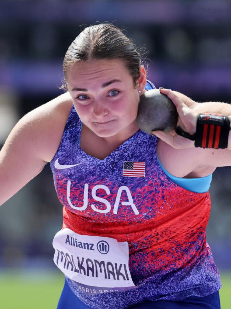 A woman in a Team USA uniform holds a shot put up by her neck to prepare to throw it