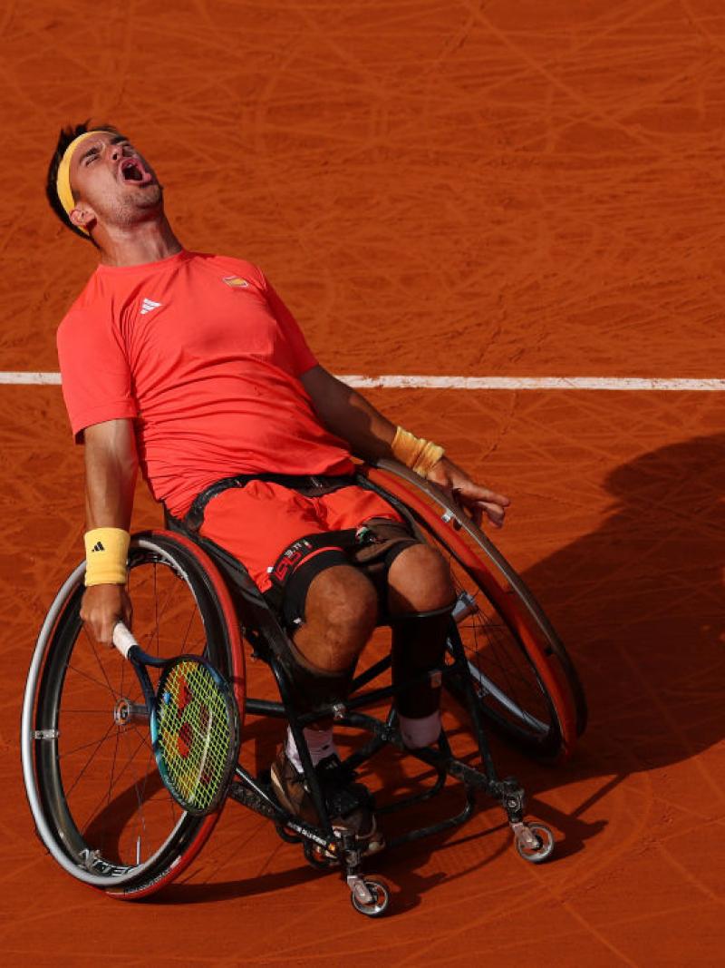 Man leans back in his wheelchair holding a tennis racket closing eyes and screaming, against a backdrop of a clay tennis court