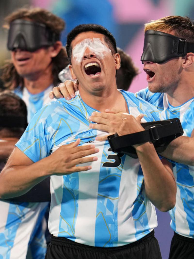 A group of men wearing Argentinian blind football uniforms and black eyeshades yell and hug each other in celebration