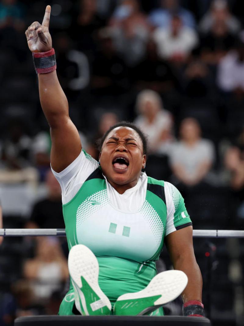 Woman in white and green sport outfit is seated with her hand in the air in a Number 1 sign, she scrunches her face to scream, with powerlifting weights behind her