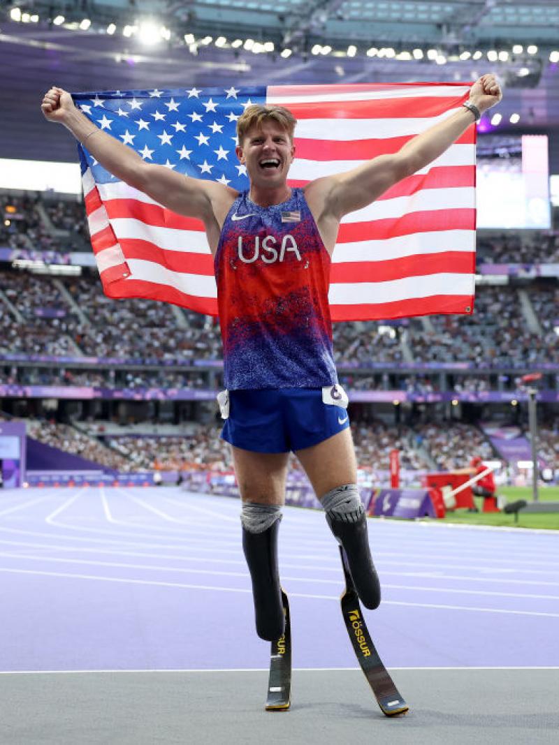 A man with running blades wearing a Team USA uniform holds up an American flag and smiles inside a full stadium