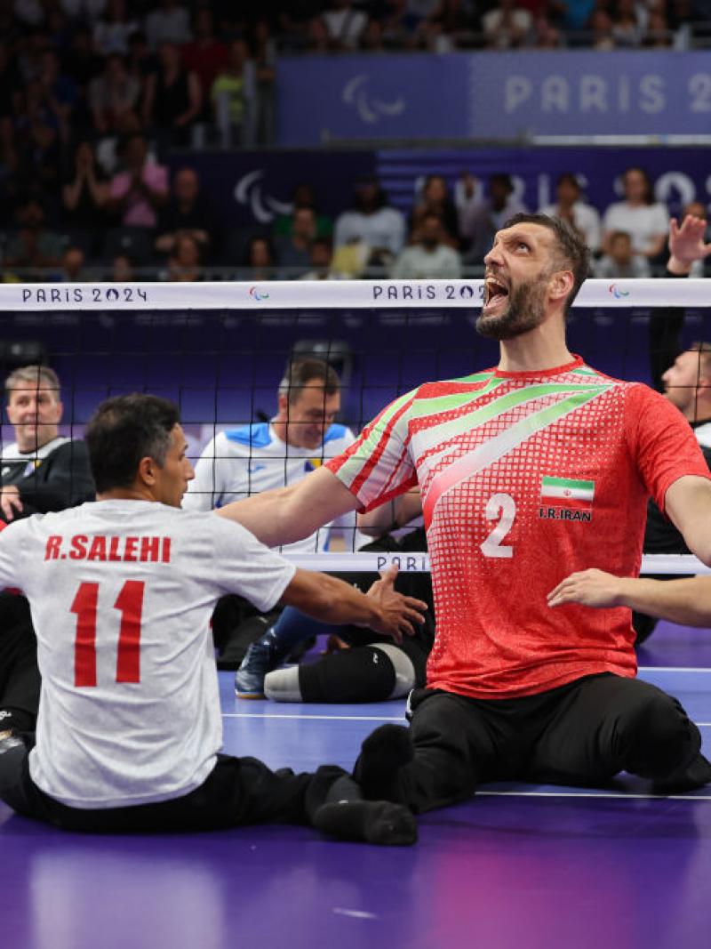 Five players wearing Team Iran uniforms yell in celebration during a sitting volleyball match