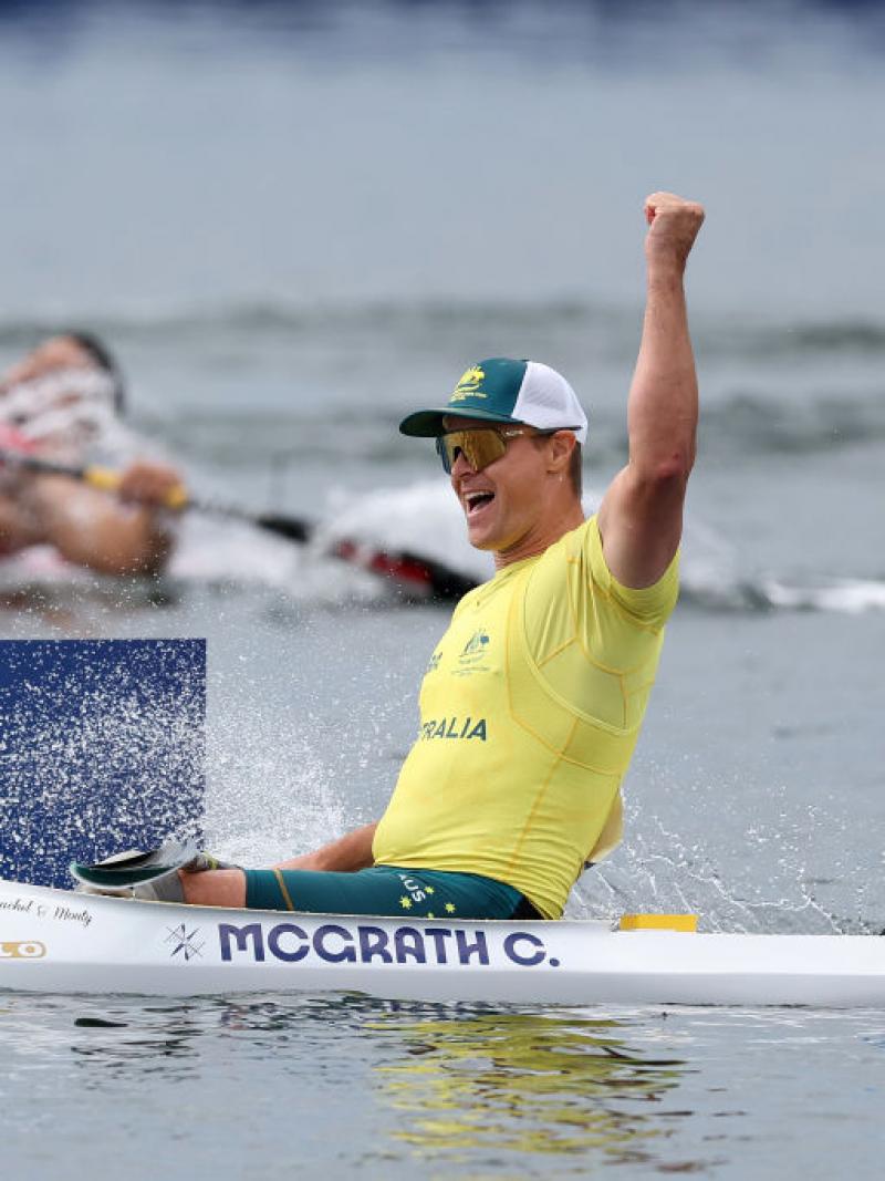 An Australian male rower wearing a yellow shirt, sunglasses and a green hat, celebrates a rowing win by punching the air in his boat