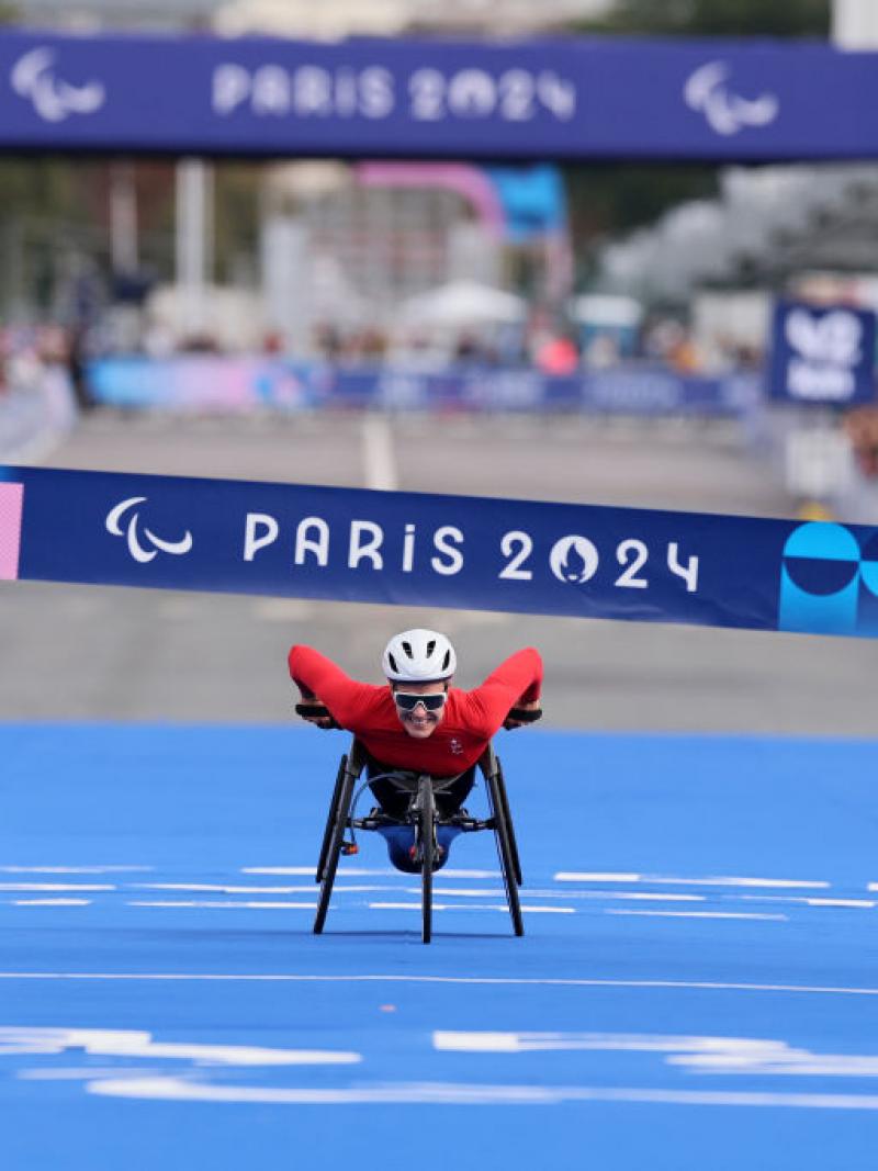 Woman in white helmet, sunglasses and red outfit leans forward in her racing wheelchair with a big smile as she approaches the finish line