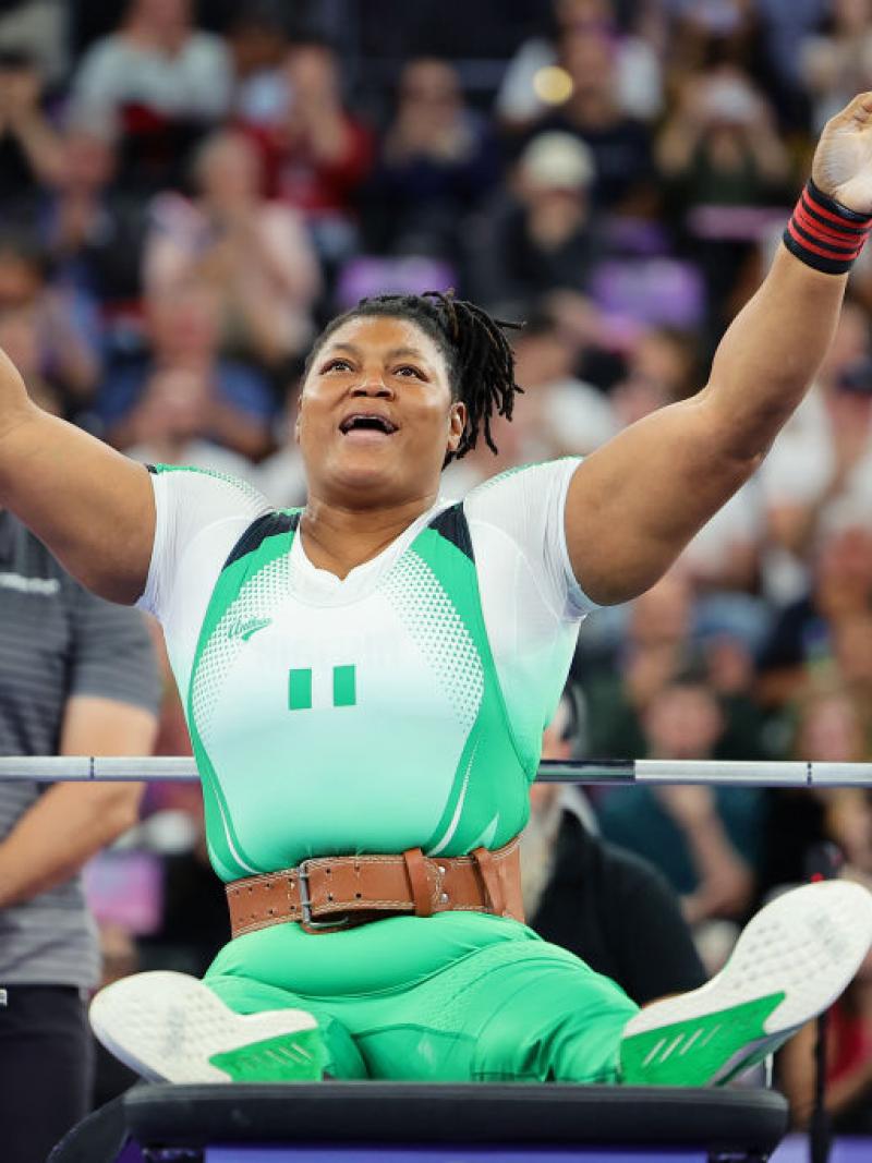 A Para powerlifter in a Team Nigeria uniform sits up on a lifting bench and raises her arms in the air in celebration