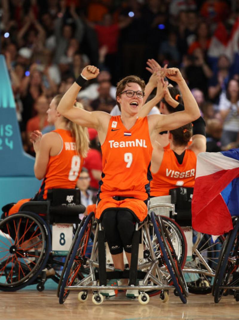 The Dutch women's wheelchair basketball team clap and raise their arms in the air, with one woman in the middle lifting up both arms and smiling