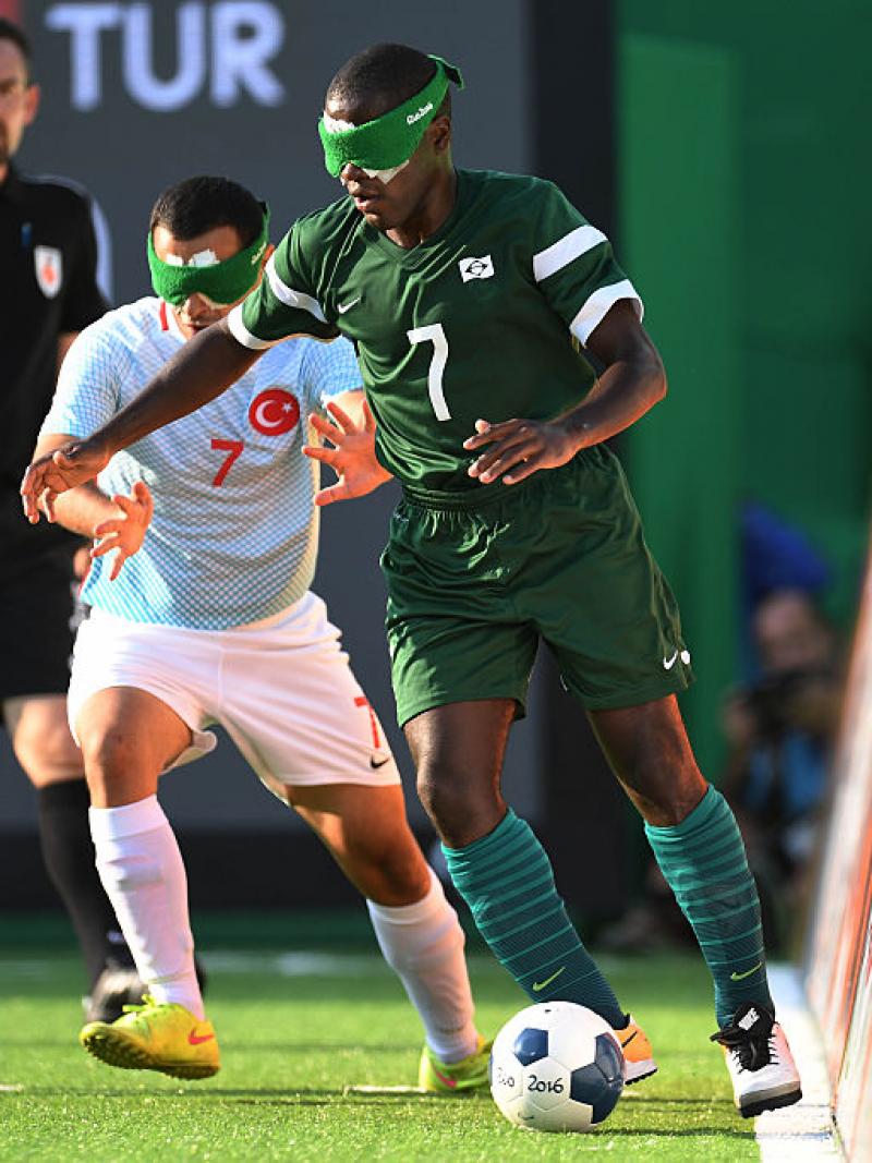 A man in green with a green blind fold tackles a man in blue taking the football away
