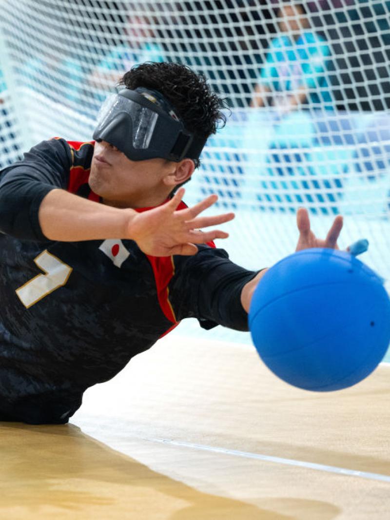 A male goalball player, wearing eye shades, is blocking a blue ball in front of the goal