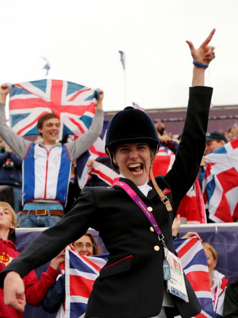 A female Para equestrian athlete poses for a photograph in front of the stands. Spectators are holding the GB flag