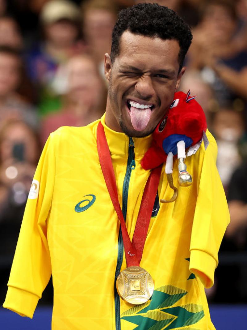 A male Para swimming athlete poses for a photo on the podium. He sticks his tongue 