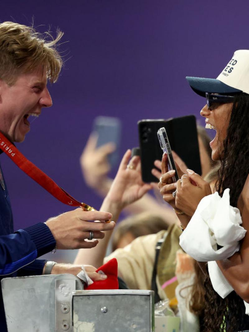 A male athlete shows his gold medal to a woman in the stands. She is holding her smartphone