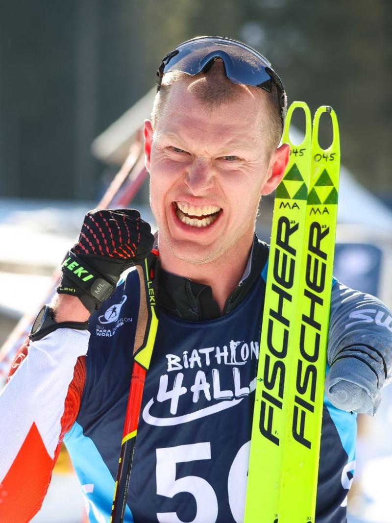 A male Para biathlon athlete smiles at the camera after competition. He is holding his skis