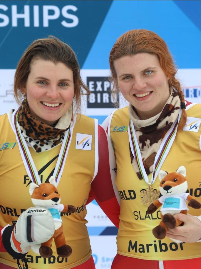 Two female athletes wearing a gold bib smile on the podium