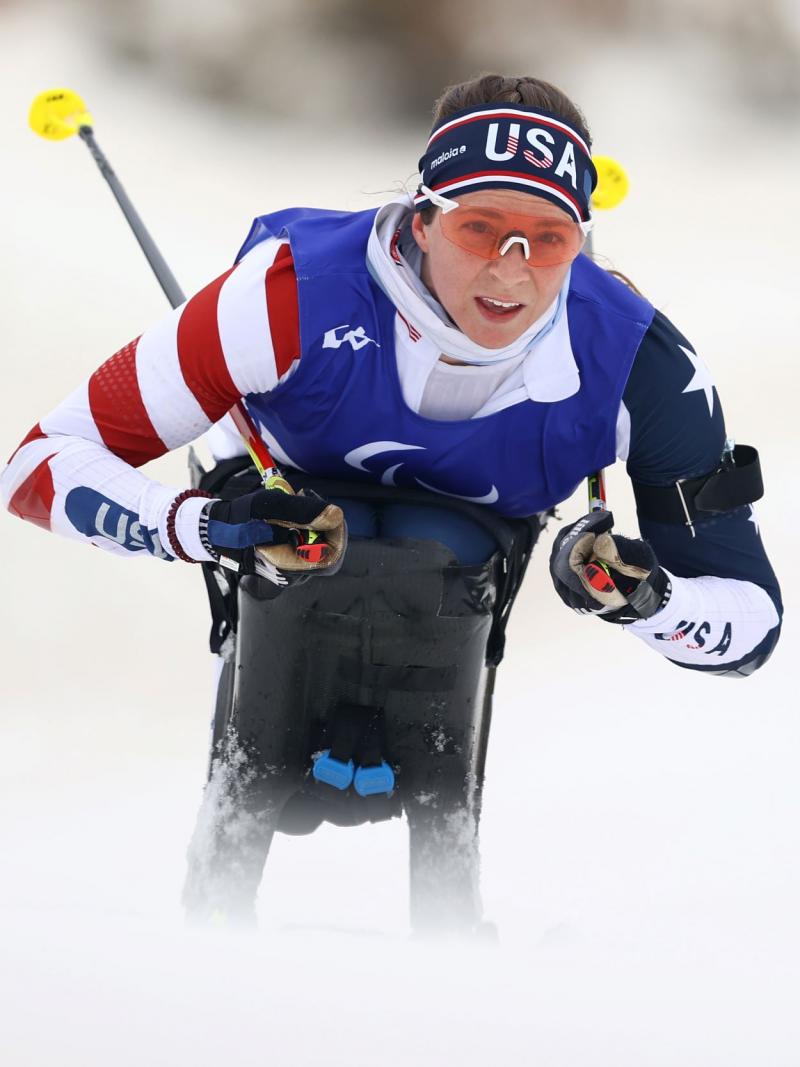 A female athlete competes in a sit-ski. She is wearing a head band that says USA and uniform in red, white and blue.