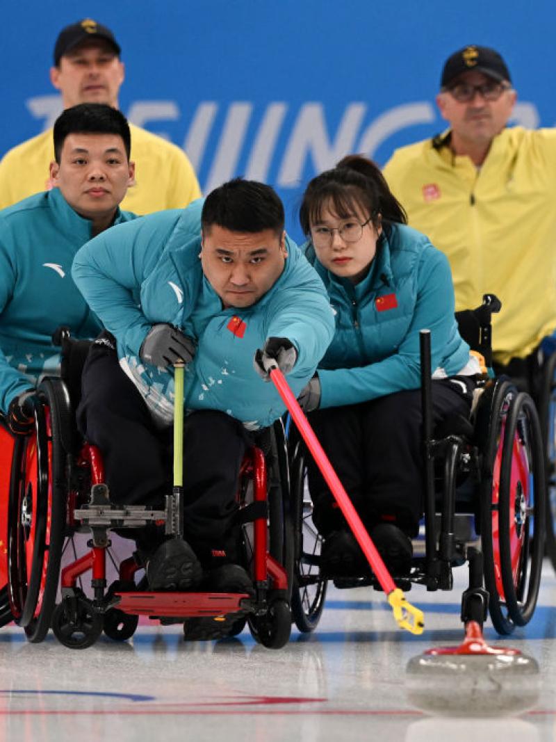 A male wheelchair curling athlete in action. He is trying to reach a curling stone with a red stick while his teammates are holding onto his wheelchair