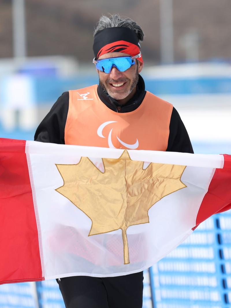 A male athlete wearing sunglasses and an Orange bib holds the Canadian flag