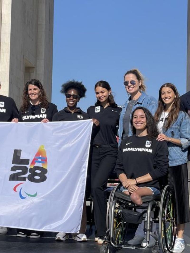 11 people including one female Paralympian pose for a group photo in front of a stadium. They are holding a white flag with the logos of the LA Games