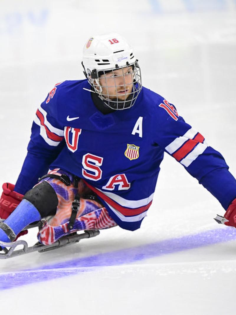 A male Para ice hockey player, who is wearing a blue Team USA kit and helmet, is in action