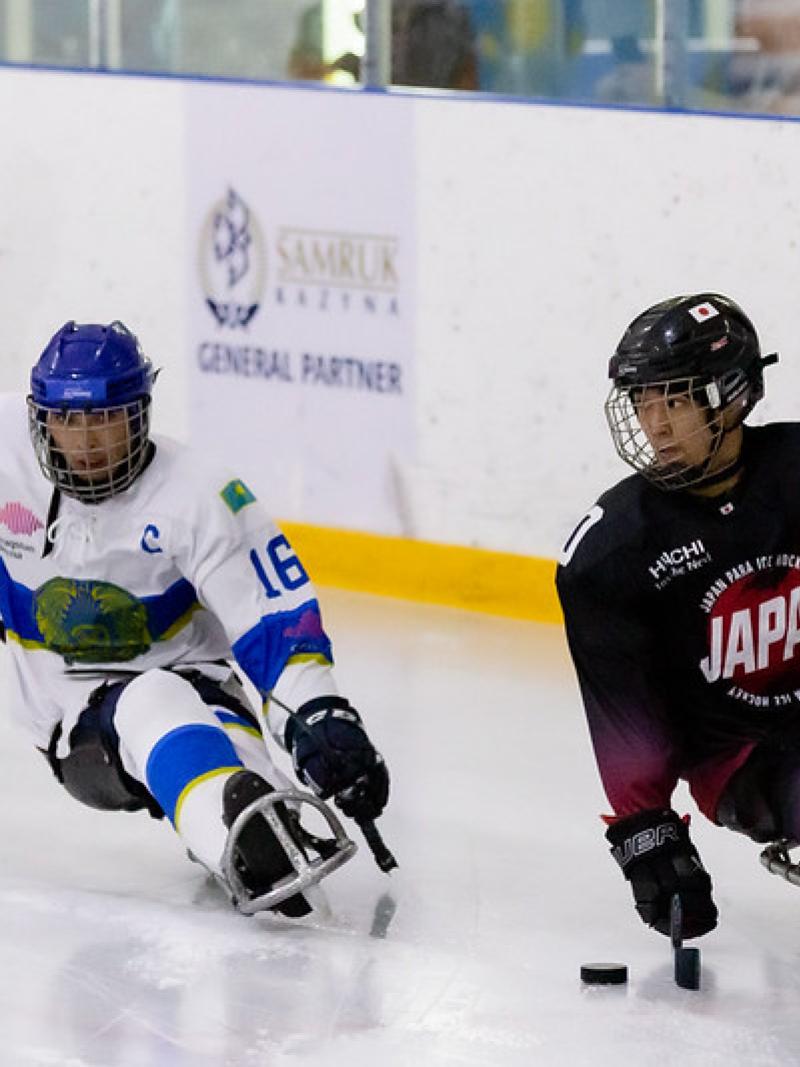A Japanese Para ice hockey player with the puck with a Kazak player behind him