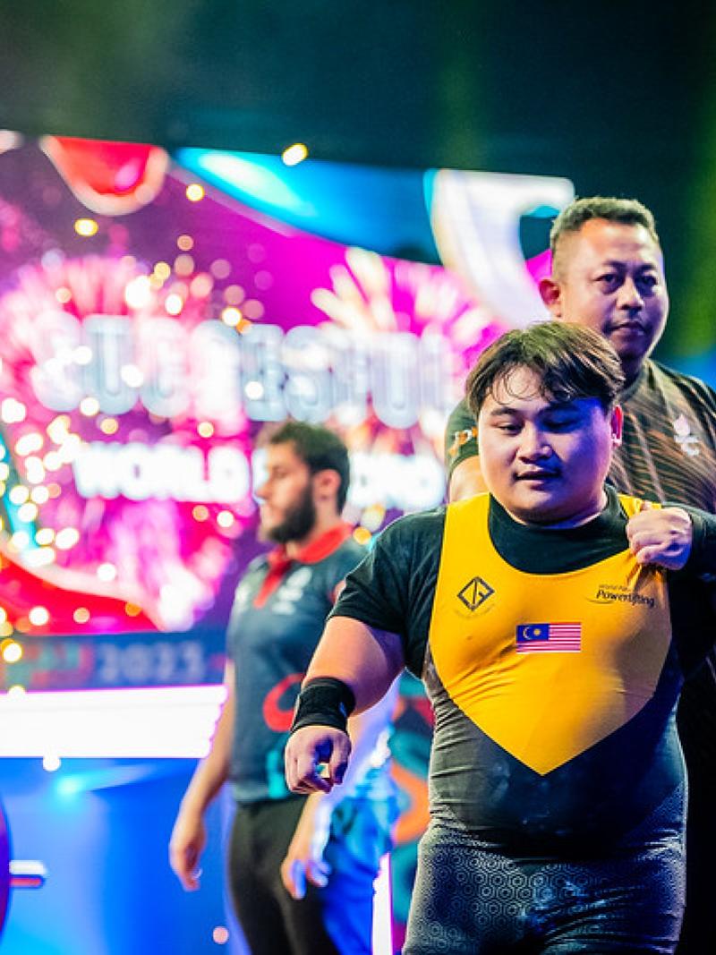 A short stature Para powerlifter on stage with three people in the background