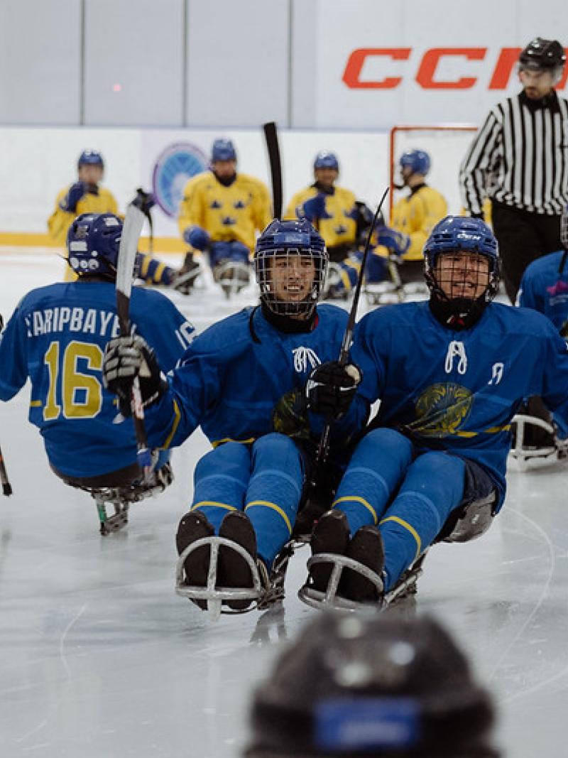 A group of four Kazak Para ice hockey players on ice with an official and four other players in the background