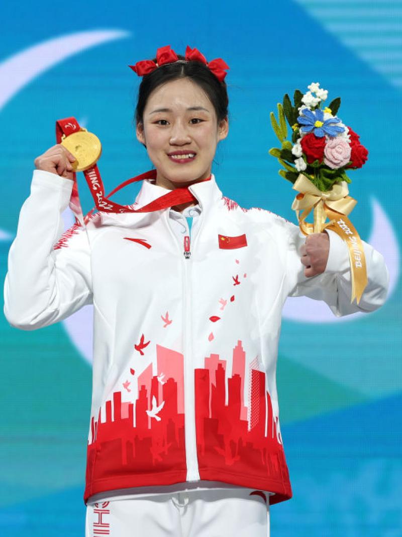 A female athlete with red ribbons on her head smiles on the podium while holding a flower bouquet and a gold medal