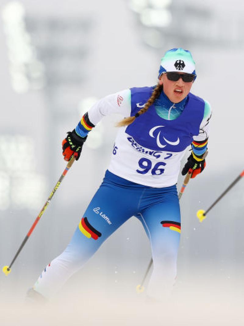 A female skier is competing behind a sighted male guide, who is looking back at her
