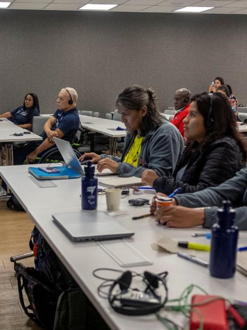 About 10 people sit in a classroom, listening to a male official speak 