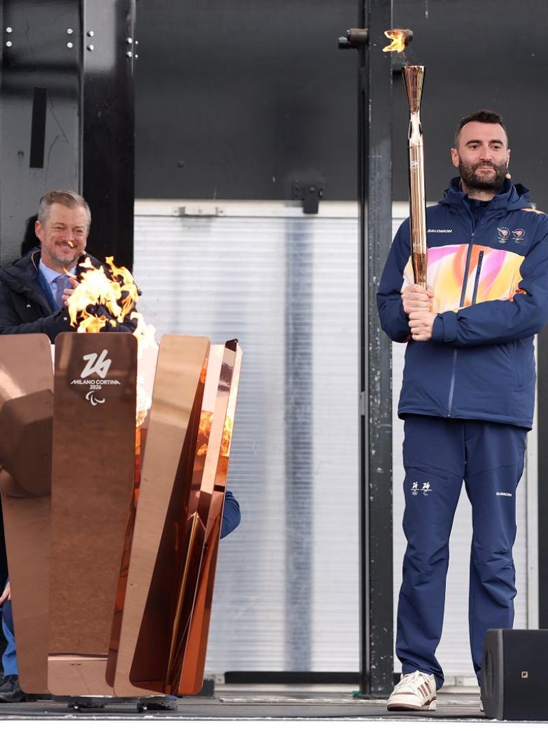 Three people are on the stage in front of a gold cauldron, with two of the people holding a gold torch.