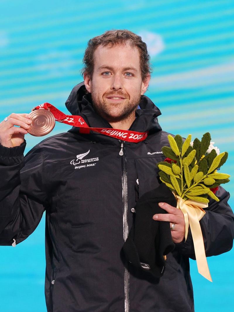 A male Para alpine skier celebrating his win, by holding a bronze medal and flowers