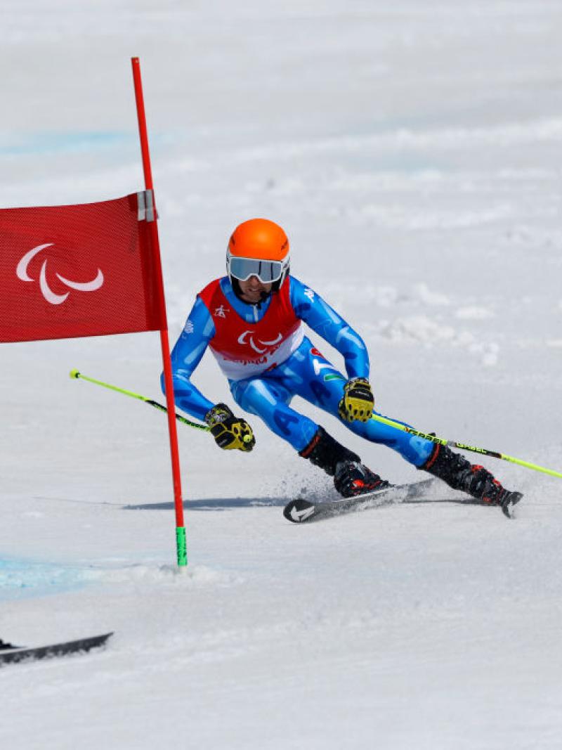 A male Paralympic alpine skiing athlete wearing a blue uniform and an orange headset is skiing behind his guide at the Beijing 2022 Paralympics.