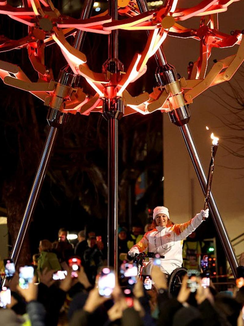 A woman is holding the Milano Cortina 2026 Paralympic torch to light a sphere-shaped cauldron in front of a crowd.