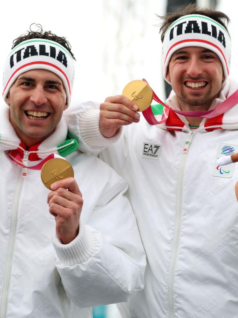 One male Para athlete and his male guide celebrating while holding their gold medals in one hand and the Milo Paralympic mascot in the other hand, wearing headbands with the words ''Italia'' and smiling at the camera