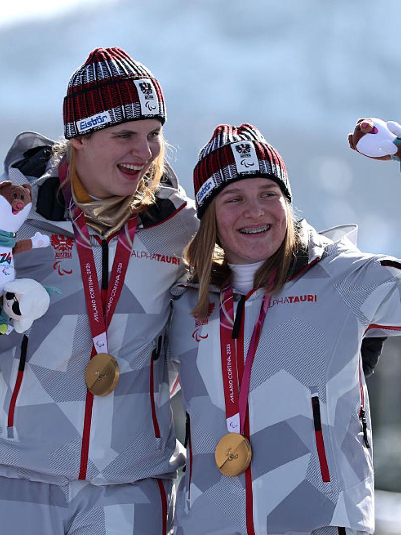 Two female Para alpine skiers celebrate on the podium after receiving their gold medals