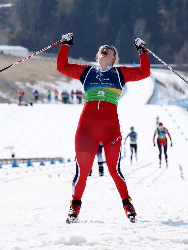 A female Para cross country skier celebrating pointing at the sky, as she crosses the finish line