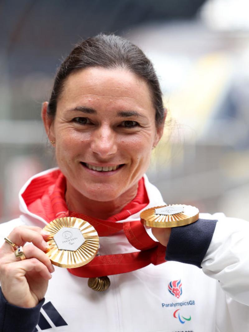 A female Para athlete wearing a white uniform is holding two gold medals in front of face and smiles