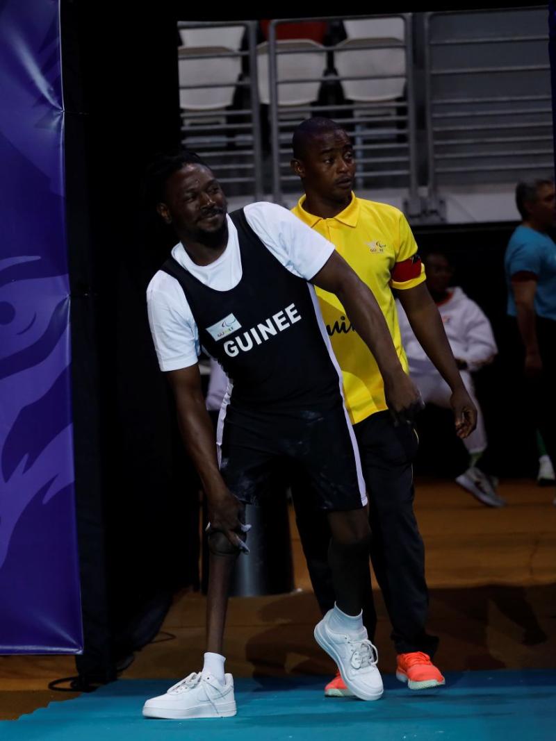 A male athlete wearing a black uniform saying Guinee walks into a venue, next to a banner that says African Open Championships