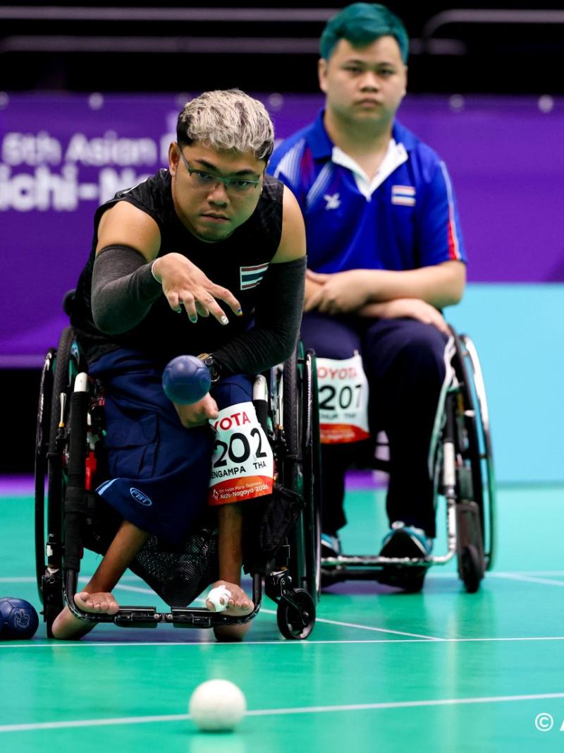 A male athlete, who is in a wheelchair, is throwing a blue boccia ball on the court