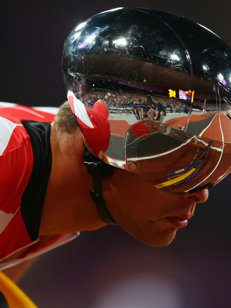 A man with a silver helmet and red jersey leans forward on his racing wheelchair
