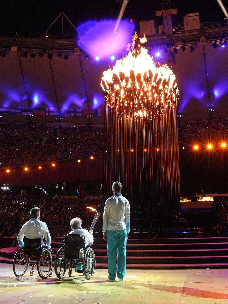 A picture of one man and woman in wheelchairs and one man standing watching a lightened cauldron during a ceremony