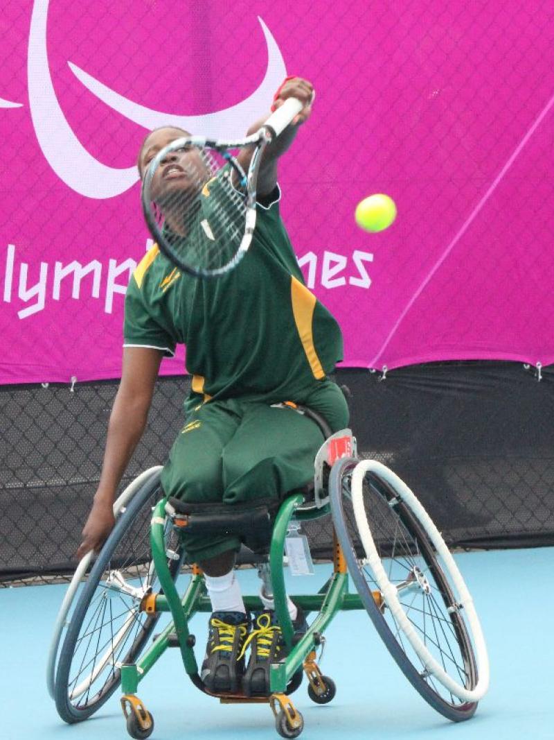 A picture of a black woman in a wheelchair serving during a wheelchair tennis match.