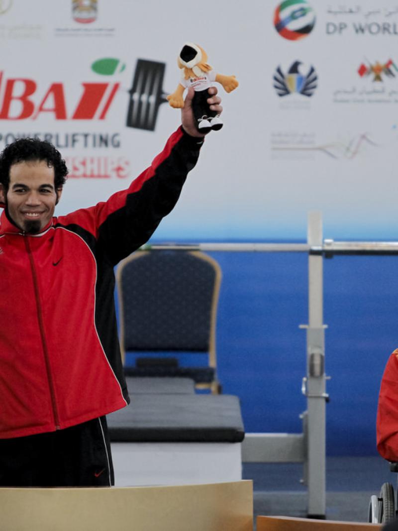 An athlete shows off his medal to a crowd, holding up a stuffed animal mascot with the other hand.