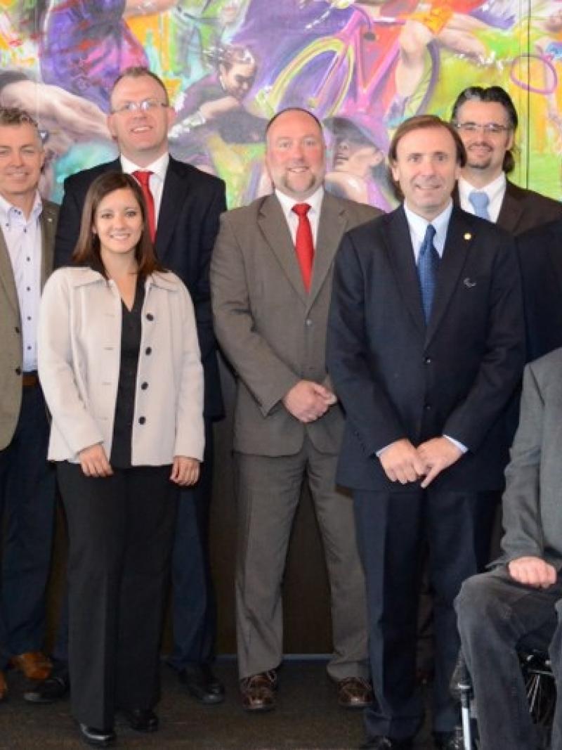 A group shot of 11 smartly dressed people lined up in front of a painted sport wall mosaic looking directly at the camera