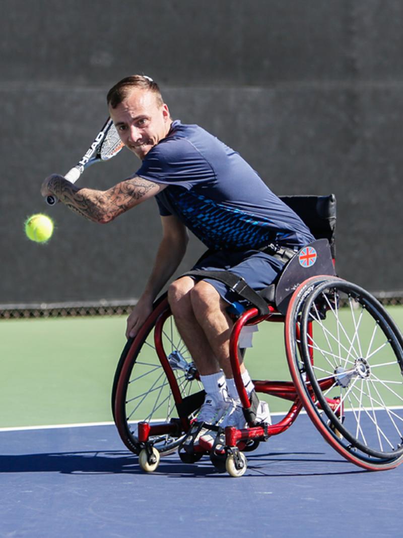 A man in a wheelchair swings back a tennis racket in preparation of hitting a backhand.
