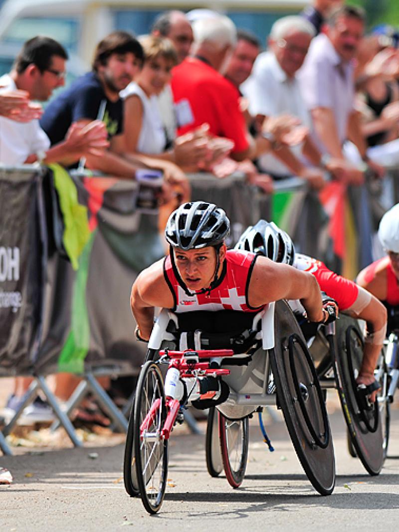 A woman looks up as she races in a wheelchair at the front of the pack with fans along the side of the road.