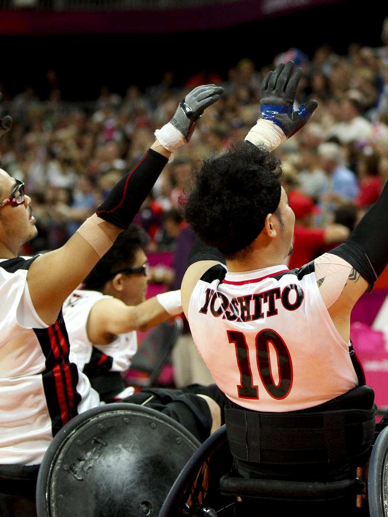Wheelchair rugby players shown from behind raise their arms and wave to a crowd.