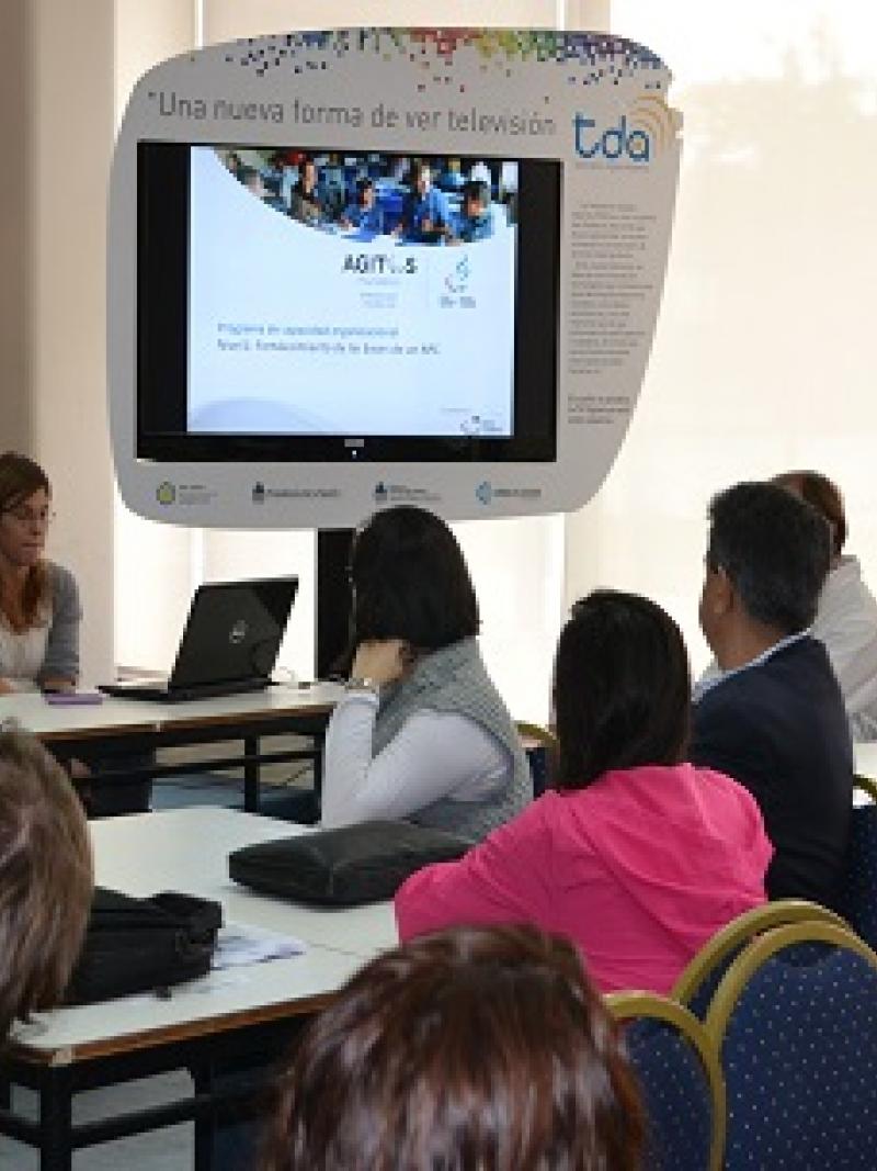 People sitting on chairs around tables, looking at a screen. Classroom atmosphere